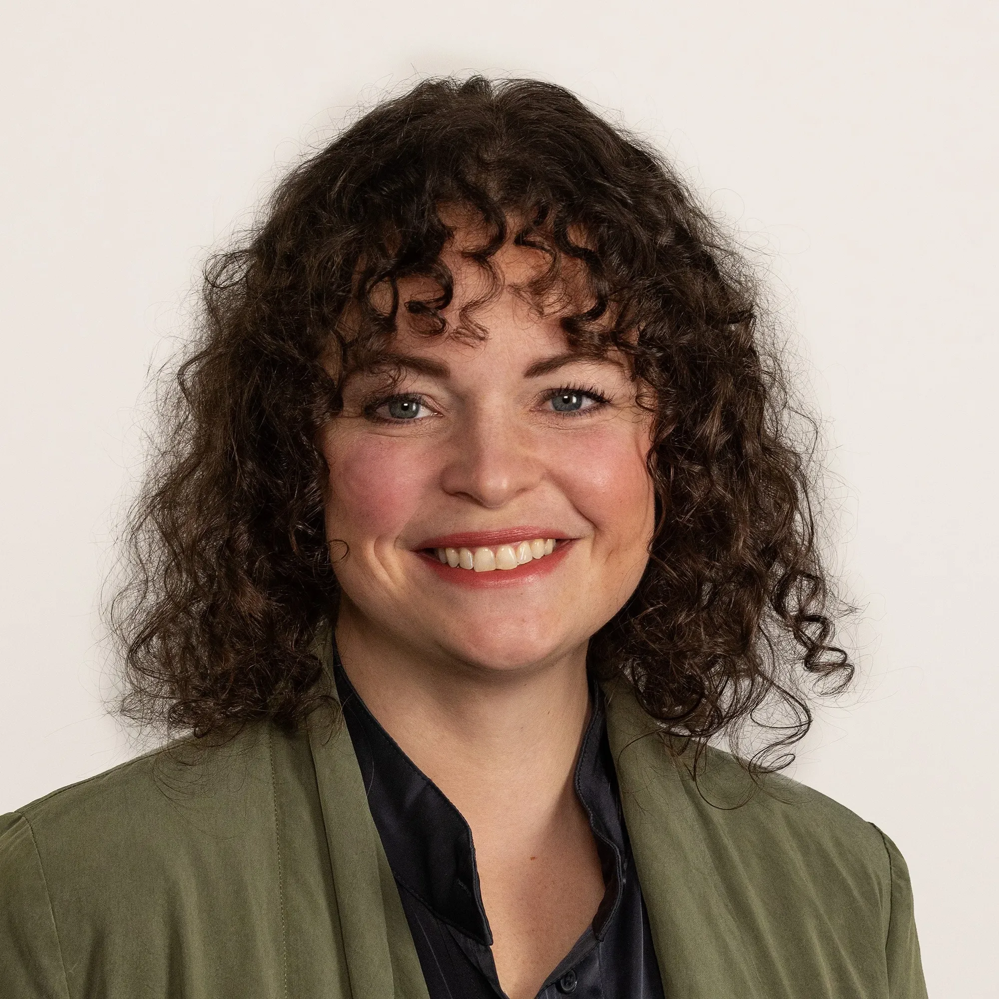 A smiling woman with shoulder-length curly brown hair wearing an olive green jacket over a black collared shirt, against a plain white background.
