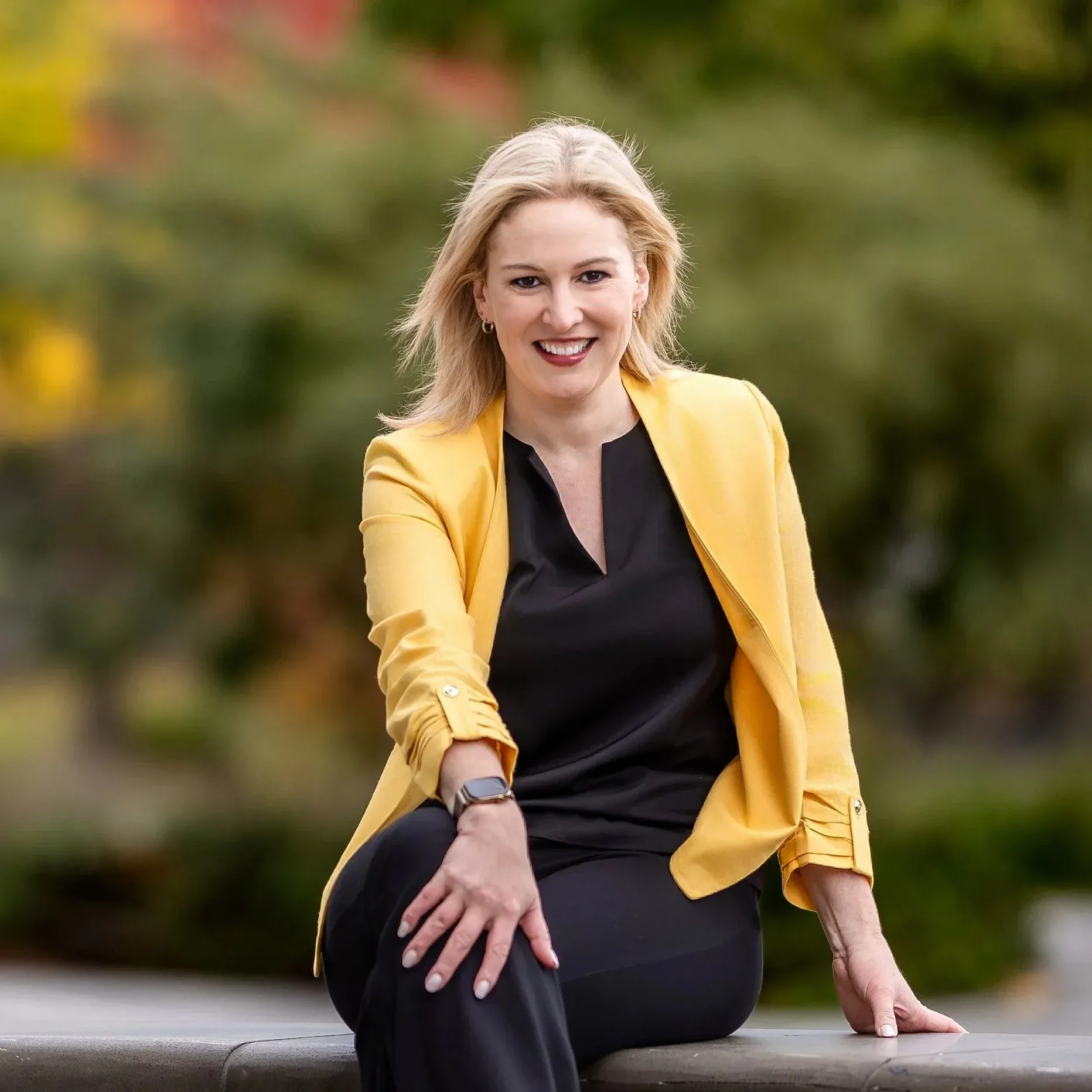 A woman with blonde hair smiling, wearing a yellow blazer and black top, kneeling outdoors with a blurred green foliage background.