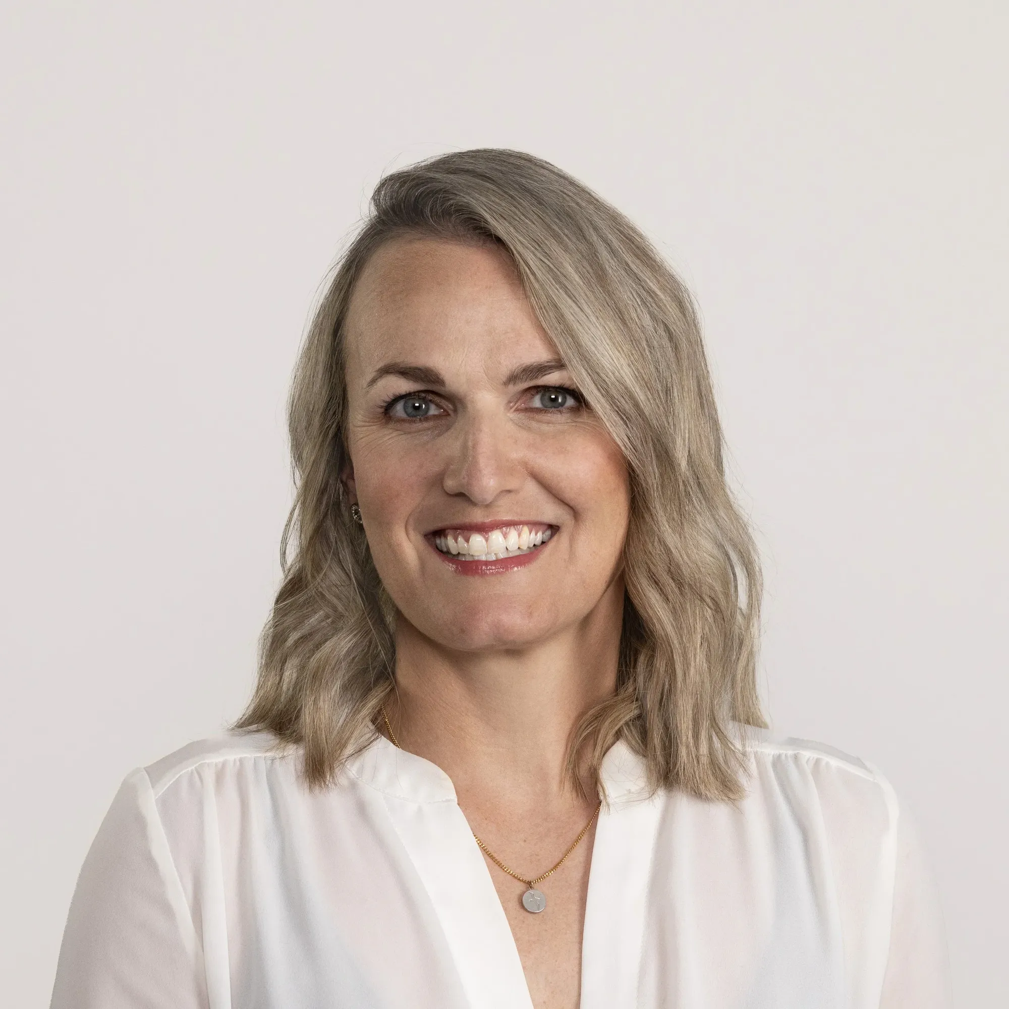 A smiling woman with shoulder-length blonde hair, wearing a white blouse and a gold necklace, against a plain white background.